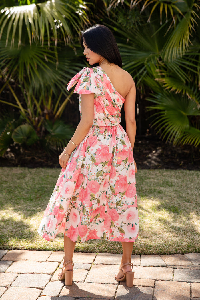 Woman wearing a pink floral dress standing in front of green foliage.