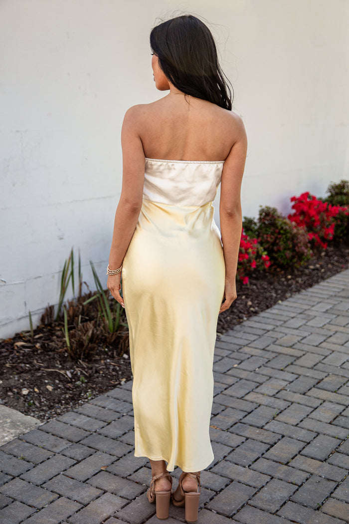 Woman in a strapless white dress standing on a paved path with a white wall and red flowers in the background.