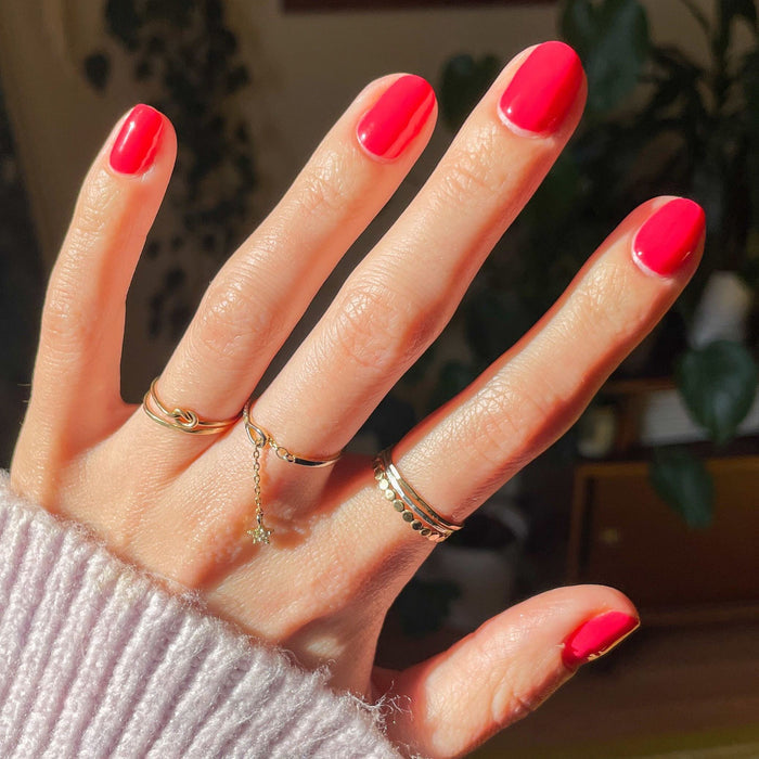 Hand with Pomodoro nail polish in vibrant pinky-tomato red on natural nails adorned with delicate gold rings