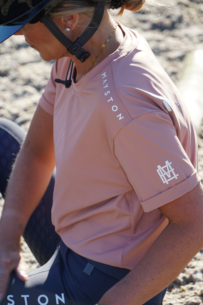 Person wearing a pink with 'Mayston' branding on a blurred outdoor background in a horse arena. luxury quality pink blush t shirt and fabric is specially crafted for active duty, providing flexibility and ease of movement for any busy lifestyle. An ideal choice for a day at the yard, in the saddle, or during your grooming duties, its loose-fitting design guarantees complete freedom. high gloss silicone branding