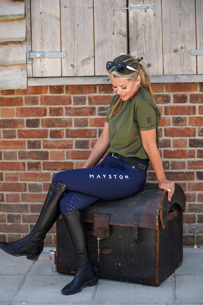Woman in equestrian attire sitting on a suitcase against a brick wall. luxury quality t shirt and fabric is specially crafted for active duty, providing flexibility and ease of movement for any busy lifestyle. An ideal choice for a day at the yard, in the saddle, or during your grooming duties, its loose-fitting design guarantees complete freedom. Hunter green colour