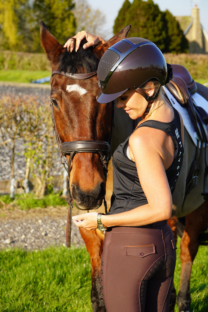 Woman in equestrian gear petting a horse by a stone wall and grass. Radipole Racerback Vest, crafted from lightweight, high-quality sports material for effortless comfort and performance. Designed to move with you, it offers breathability and freedom of motion, making it perfect for riding, training, or everyday wear. Finished with bold white high-gloss Mayston silicone branding on the chest and a sleek logo at the rear, this black vest combines sporty style with signature Mayston.