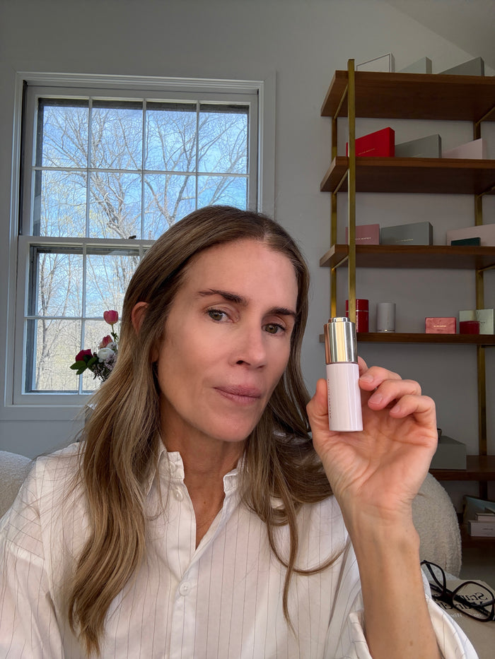 Woman holding a skincare product in a room with a window and shelves.