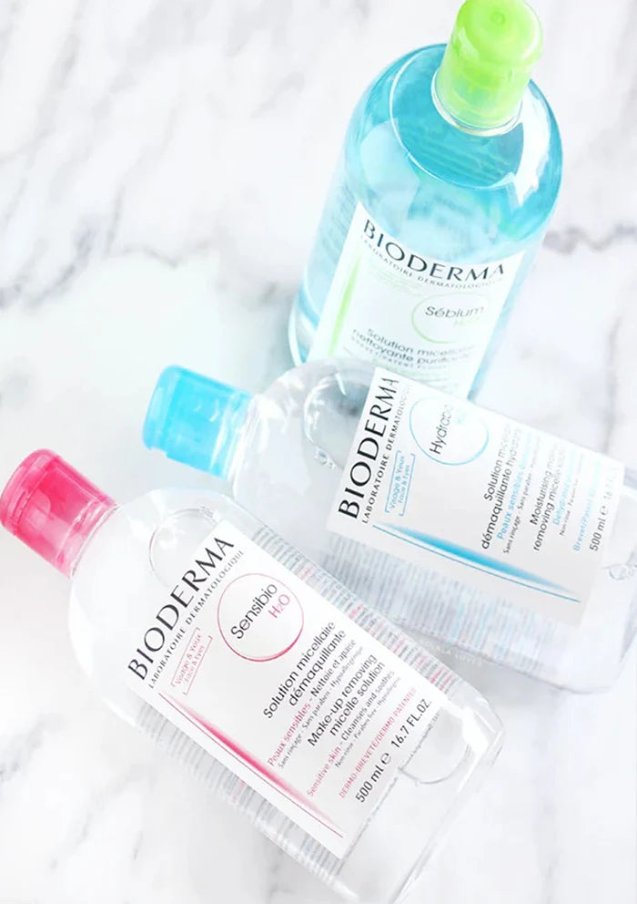 Three Bioderma skincare bottles arranged on a white marble surface: a clear bottle with a pink cap and pink text for Santé-Firming Hand Cleanser, a clear bottle with a blue cap and blue text for Hydroal Hydrating Face Cleanser, and a blue bottle with a green cap for Solisyn Blue Cleanser.