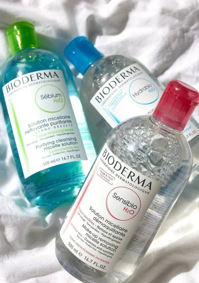 Three Bioderma facial cleansing products arranged on a white textured fabric background: Sébium H2O in a translucent blue bottle with a green cap, Hyaluve H2O in a translucent blue bottle with a blue cap, and Sensibio H2O in a clear bottle with a red cap.