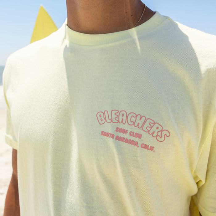 Adult model wearing yellow graphic tee with "Bleachers Surf Club" design