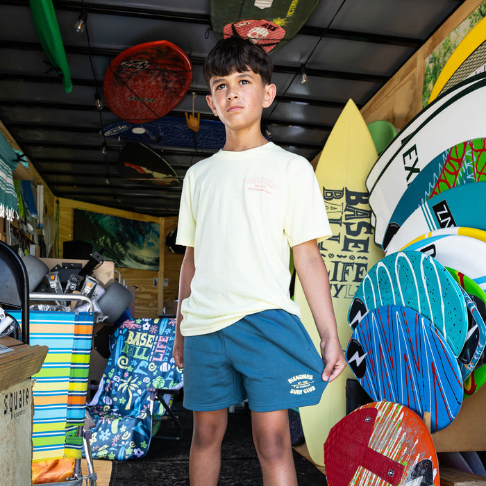 Youth model wearing blue youth shorts with "Bleachers Surf Club" design