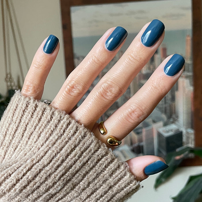 Hand with Bodysuit gel polish manicure in dusty cool teal on medium-length nails against blurred cityscape background