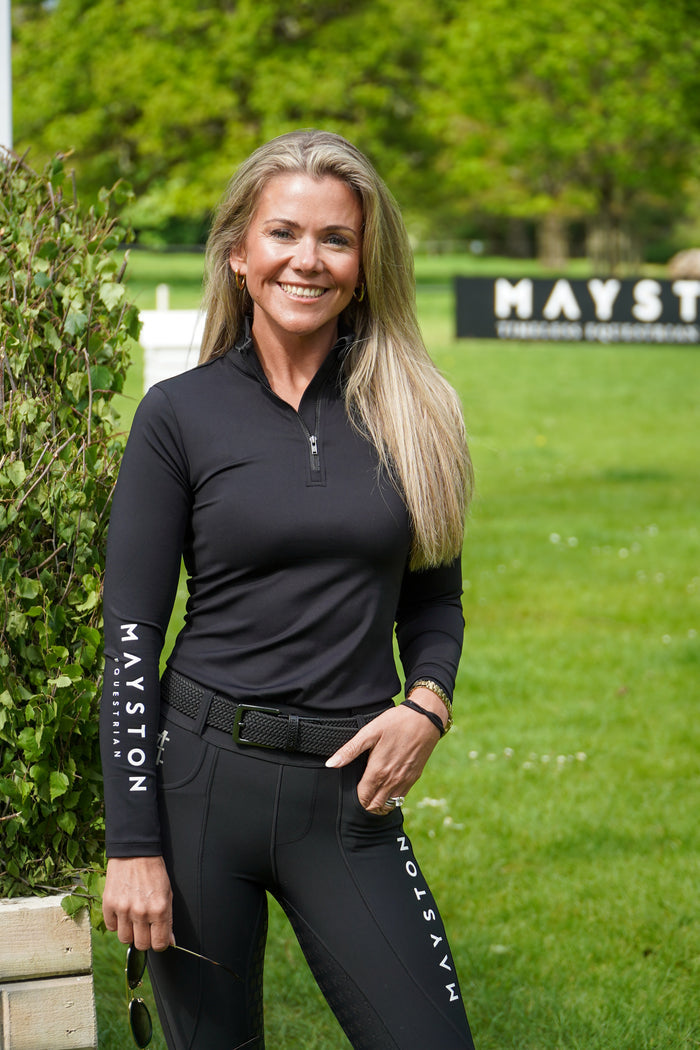 Woman wearing black equestrian attire with 'Mayston' branding in a grassy outdoor setting. Woman in equestrian attire standing in a grassy field with a Mayston banner in the background. Equestrian base layer, luxury sports material. stretch, subtle white high gloss silicone branding, 1/4 zip, colour match embroidery. Long Sleeve black base layer with  branding, woman stood wearing it on cross country course