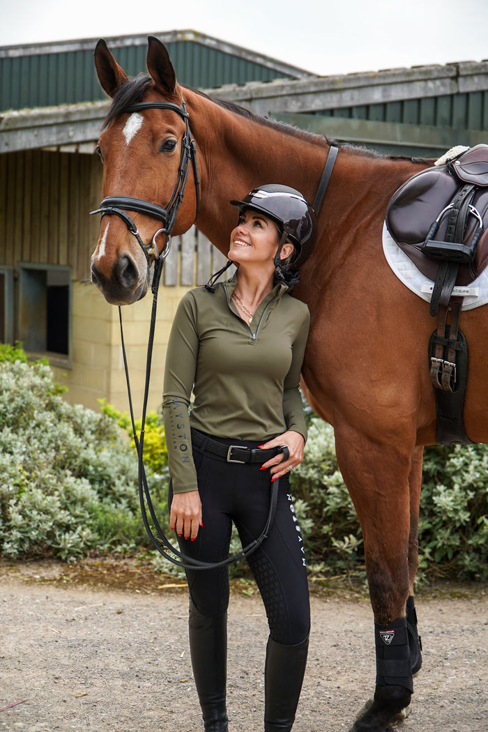 Woman in equestrian attire standing next to a brown horse in an outdoor setting. Broadstone long sleeve Base, everyday technical layer, it offers a flattering, easy silhouette that moves with you while maintaining structure and confidence. Soft, breathable and designed for performance. Country Green and detailed with sleek MAYSTON branding along the sleeve and collar