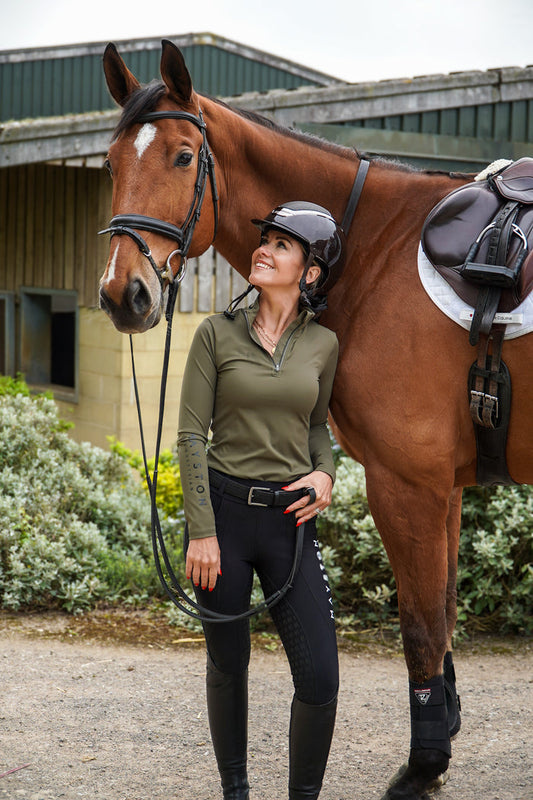Woman in equestrian attire standing next to a brown horse in an outdoor setting. Broadstone long sleeve Base, everyday technical layer, it offers a flattering, easy silhouette that moves with you while maintaining structure and confidence. Soft, breathable and designed for performance. Country Green and detailed with sleek MAYSTON branding along the sleeve and collar