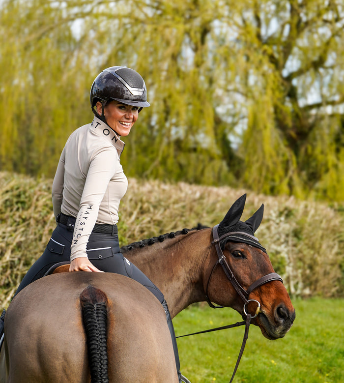 Woman riding a horse in an outdoor setting with trees in the background. Broadstone long sleeve Base, everyday technical layer, it offers a flattering, easy silhouette that moves with you while maintaining structure and confidence. Soft, breathable and designed for performance. Taupe Beige and detailed with sleek MAYSTON branding along the sleeve and collar