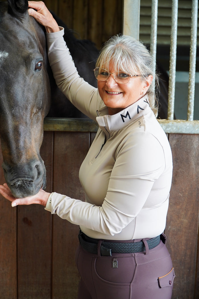 Woman in equestrian attire petting a horse in a stable. Broadstone long sleeve Base, everyday technical layer, it offers a flattering, easy silhouette that moves with you while maintaining structure and confidence. Soft, breathable and designed for performance. Taupe Beige and detailed with sleek MAYSTON branding along the sleeve and collar