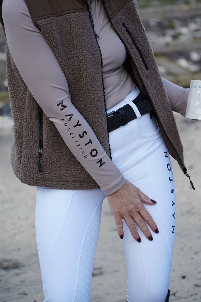 Person wearing a brown fleece vest, white breeches with 'Mayston' branding, and a black belt on a blurred outdoor background.