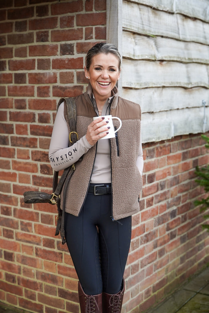 Woman holding a mug against a brick wall. Broadstone long sleeve Base, everyday technical layer, it offers a flattering, easy silhouette that moves with you while maintaining structure and confidence. Soft, breathable and designed for performance. Taupe Beige and detailed with sleek MAYSTON branding along the sleeve and collar