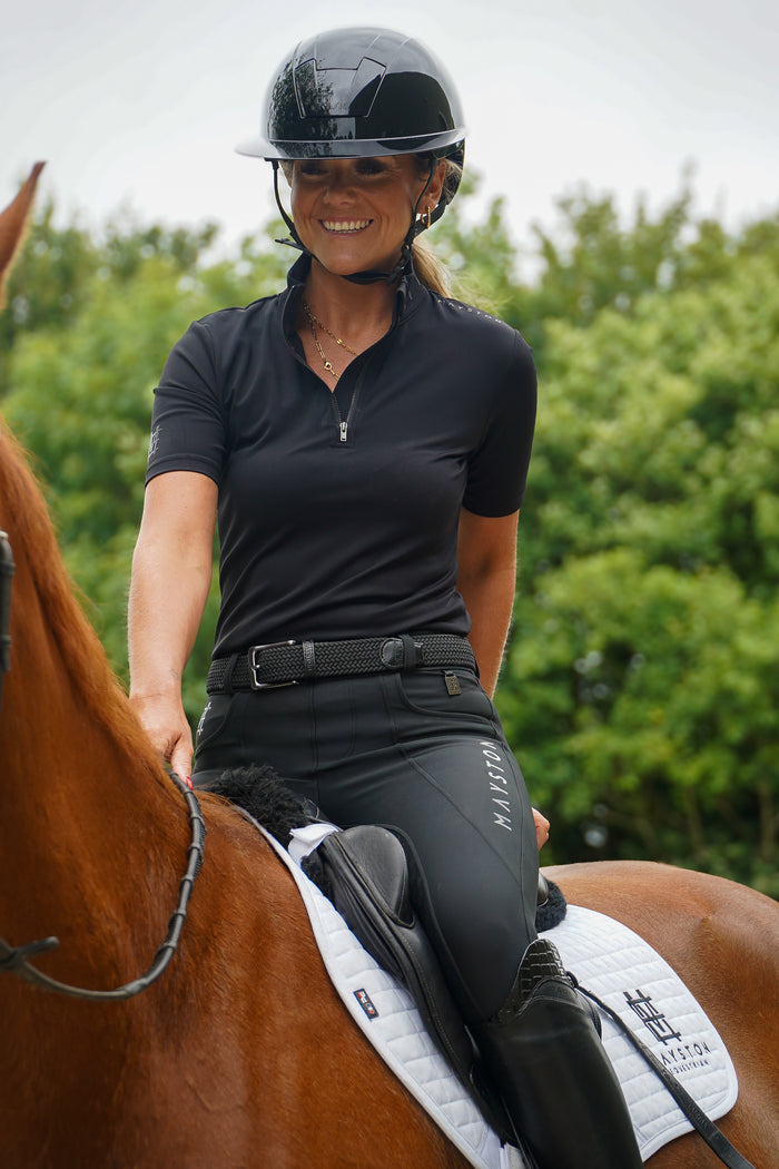 Woman riding a horse with greenery in the background. Broadstone Short sleeve Luxury Sports material base layer short sleeve. Black with subtle black branding colour match embroidery. High collar that's not restrictive. 1/4 zip. Blackout design