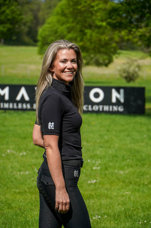 Woman in black equestrian attire standing on a grassy field with 'Mayston Equestrian' branding in the background. Broadstone Short sleeve Luxury Sports material base layer short sleeve. Black with subtle white branding colour match embroidery. High collar that's not restrictive. 1/4 zip design