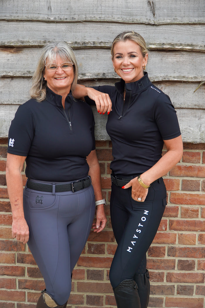Two women in equestrian attire standing against a brick wall. Broadstone Short sleeve Luxury Sports material base layer short sleeve. Black with subtle white branding colour match embroidery. High collar that's not restrictive. 1/4 zip design