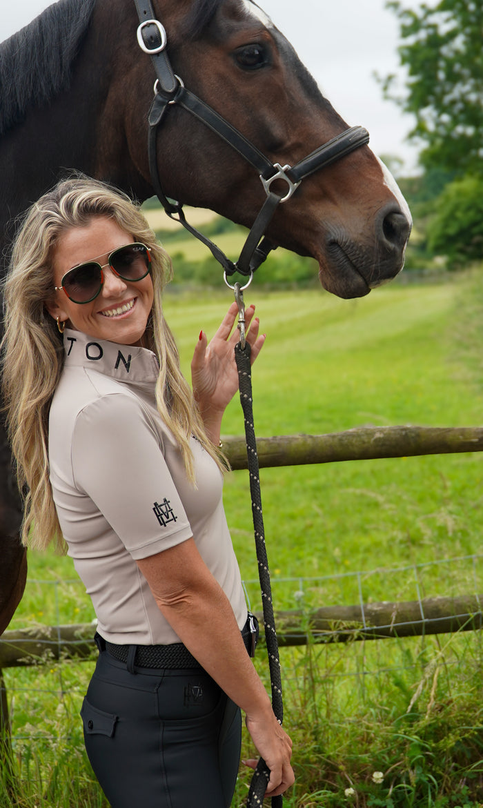 Woman in equestrian attire standing next to a horse in a grassy field.  Crafted from our soft, breathable performance fabric, it keeps you cool without compromising on style. Finished in taupe, with MAYSTON embroidery at the collar and black silicone branding on the sleeve, it’s a versatile piece that transitions effortlessly from yard to everyday wear.