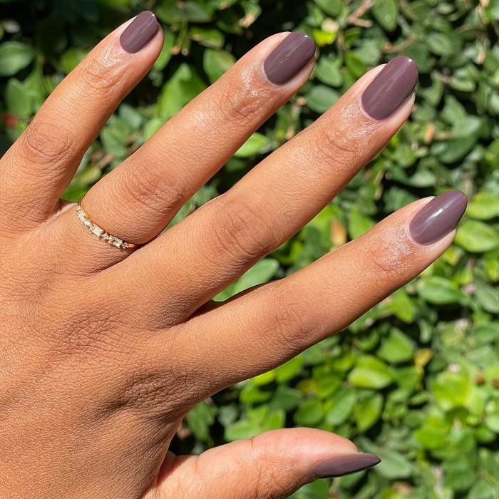 Bunny deep neutral brown gel polish on natural nails outdoors with greenery background and subtle gold ring