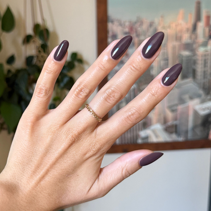 Bunny deep neutral brown gel polish on almond shaped nails with gold chain ring and cityscape background