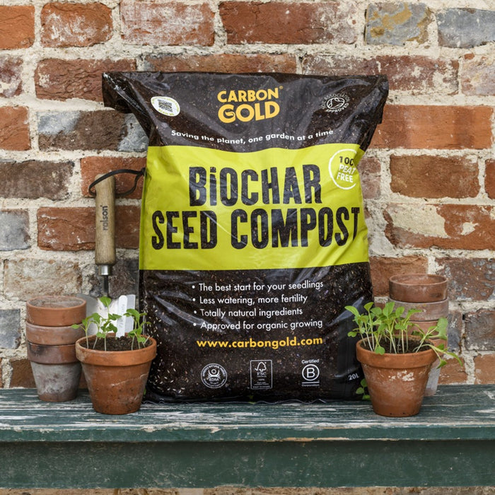 A bag of Carbon Gold Biochar Seed Compost placed against a brick wall with several small potted plants beside it.