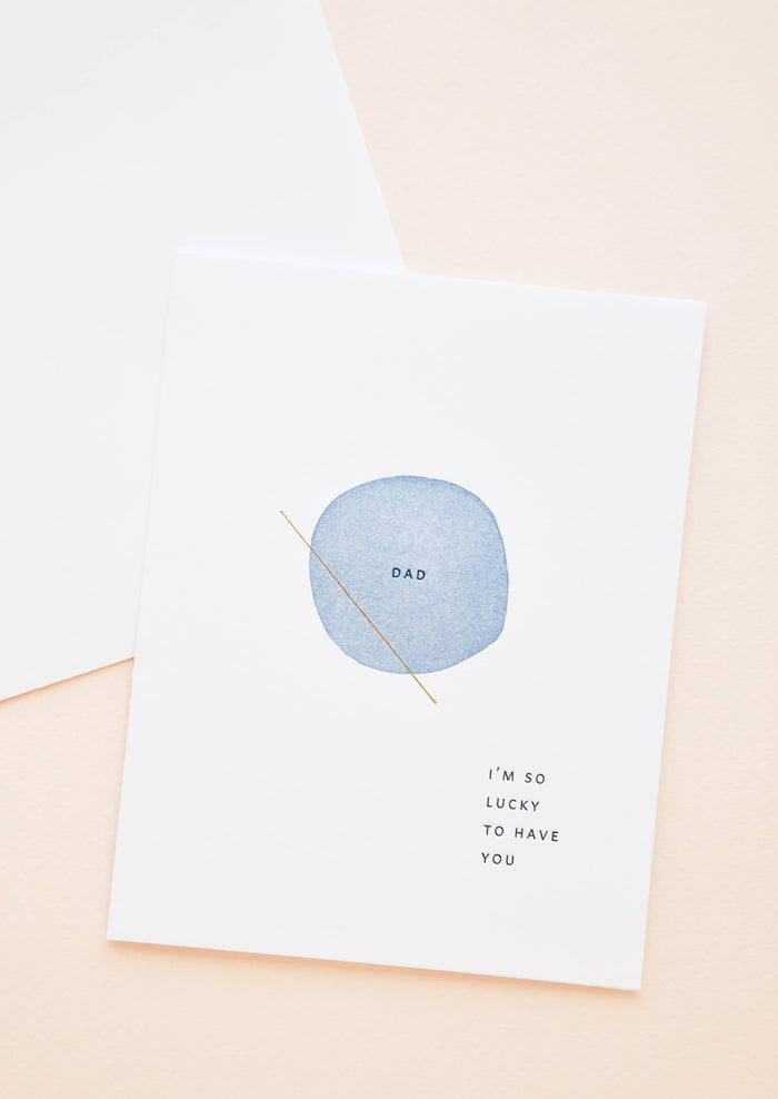 White greeting card with letter press printed blue circle at center reading "Dad". Bottom corner reads "I'm So Lucky To Have You".