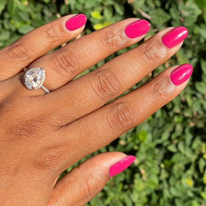 Hand with natural nails painted in vibrant Darling quick dry gel polish hot magenta against green foliage background