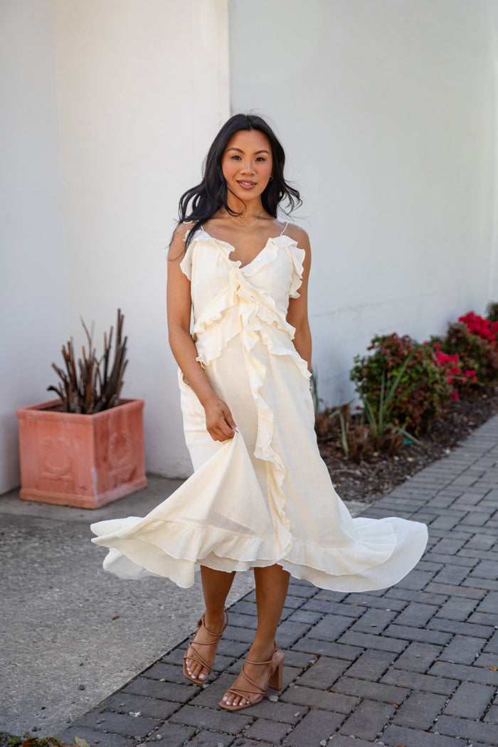 Woman in a white ruffled dress standing outdoors next to a white wall and potted plant.