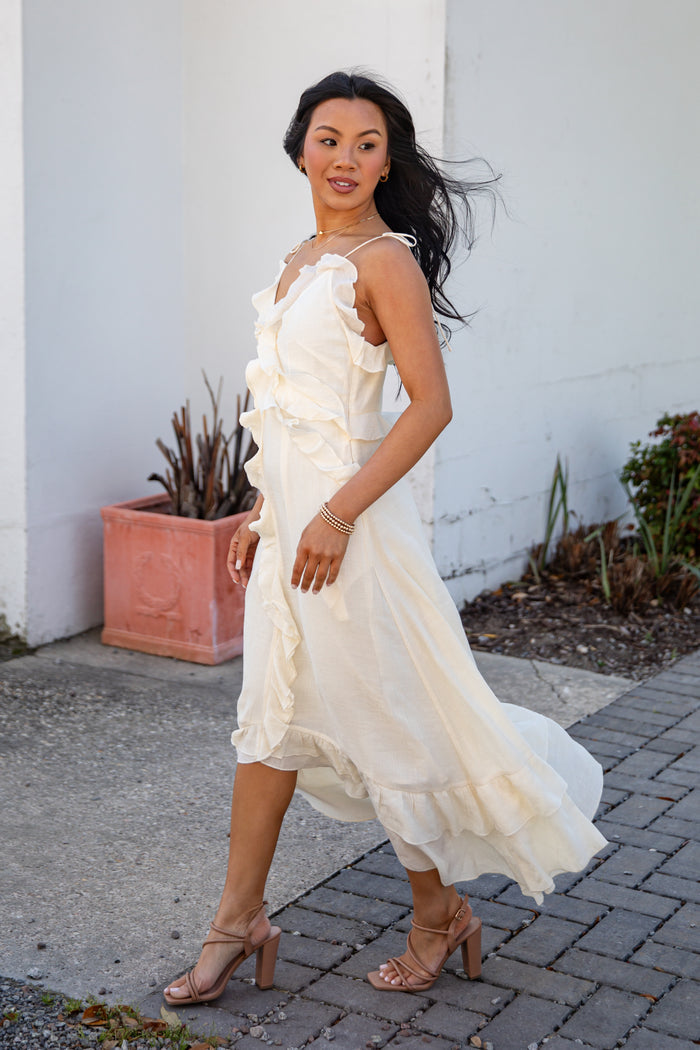Woman in a white dress standing outdoors against a white wall with plants.