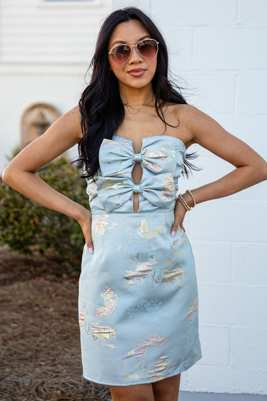 Woman wearing a light blue dress with metallic accents against a white wall.