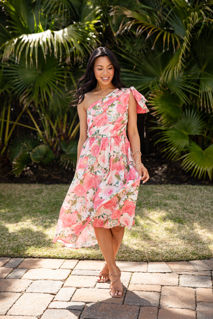 Woman in a floral one-shoulder dress standing outdoors with greenery in the background