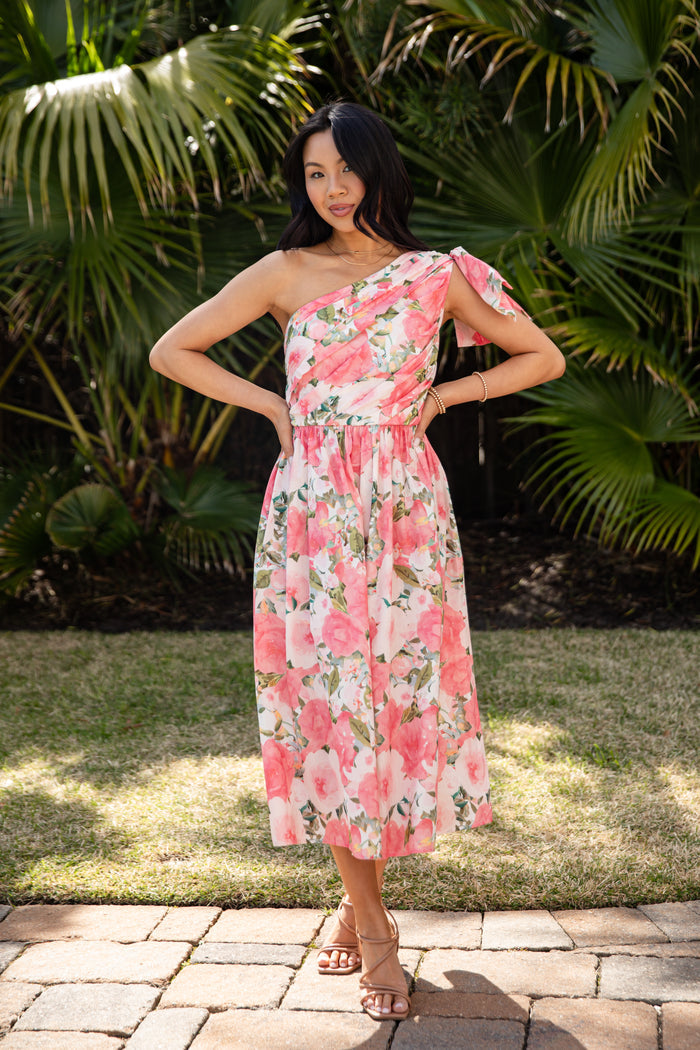 Woman wearing a floral one-shoulder dress standing outdoors with greenery in the background