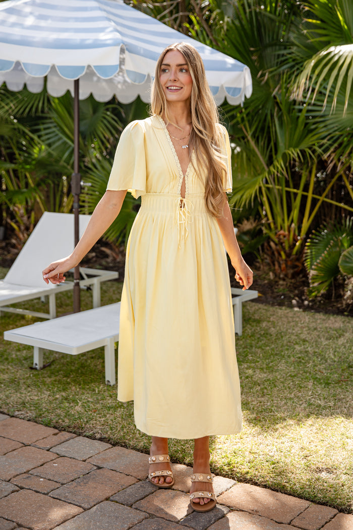 Woman in a yellow dress standing outdoors under a striped umbrella with greenery in the background.
