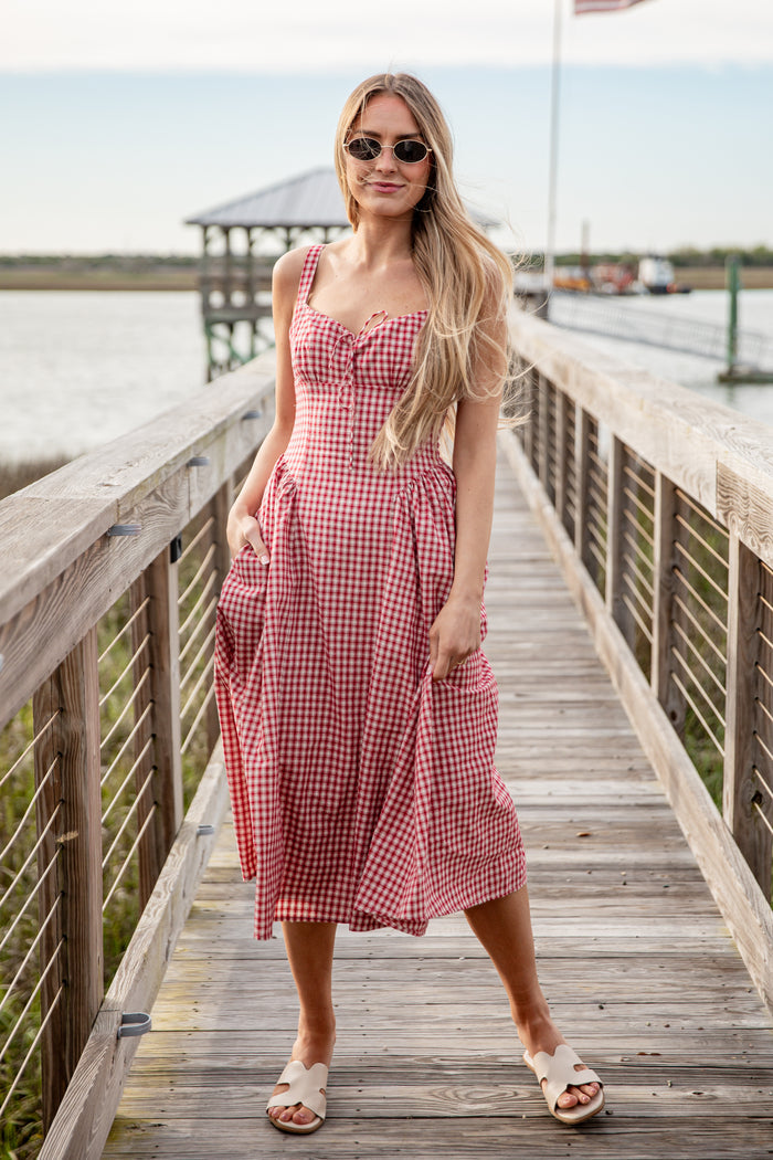 Woman in a red and white checkered dress standing on a wooden dock by a body of water.