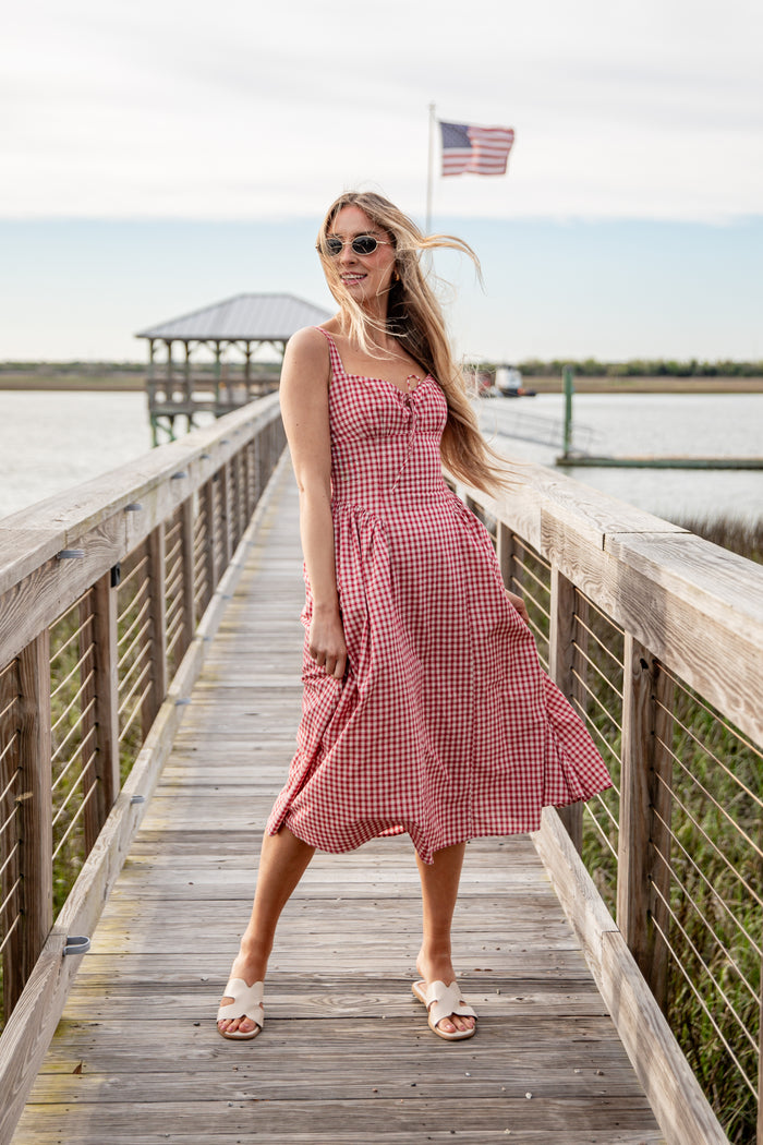 Woman in a pink checkered dress standing on a wooden dock by a lake.
