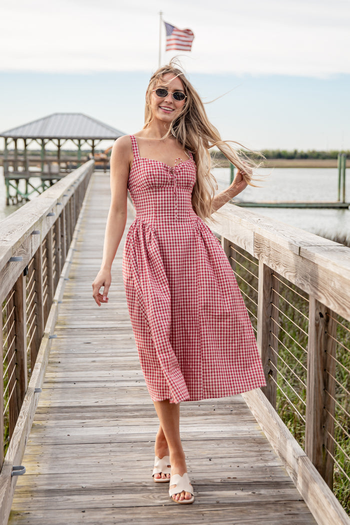 Woman in a pink checkered dress standing on a wooden dock with an American flag in the background.