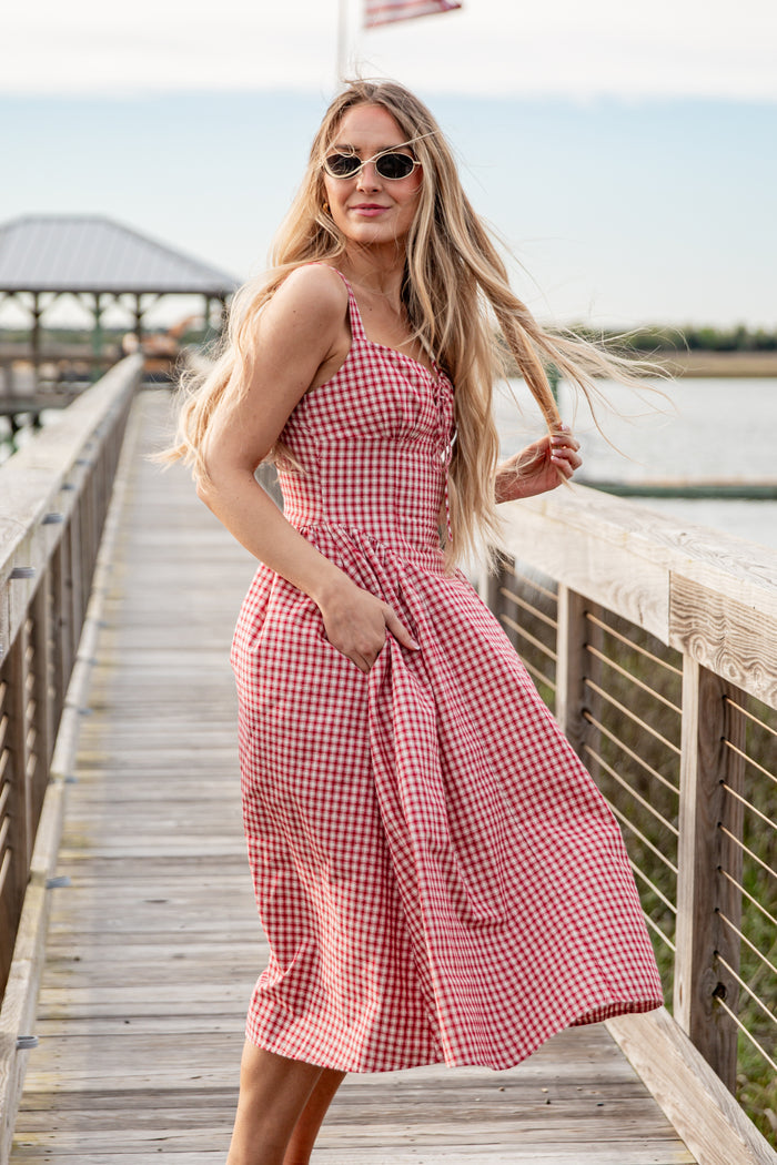 Woman in a red checkered dress standing on a wooden pier by a body of water.