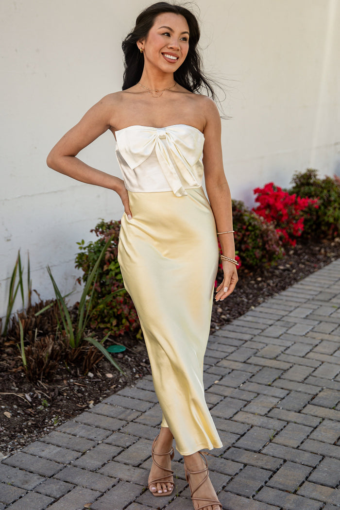 Woman in a strapless yellow dress standing on a paved path with plants and flowers in the background.