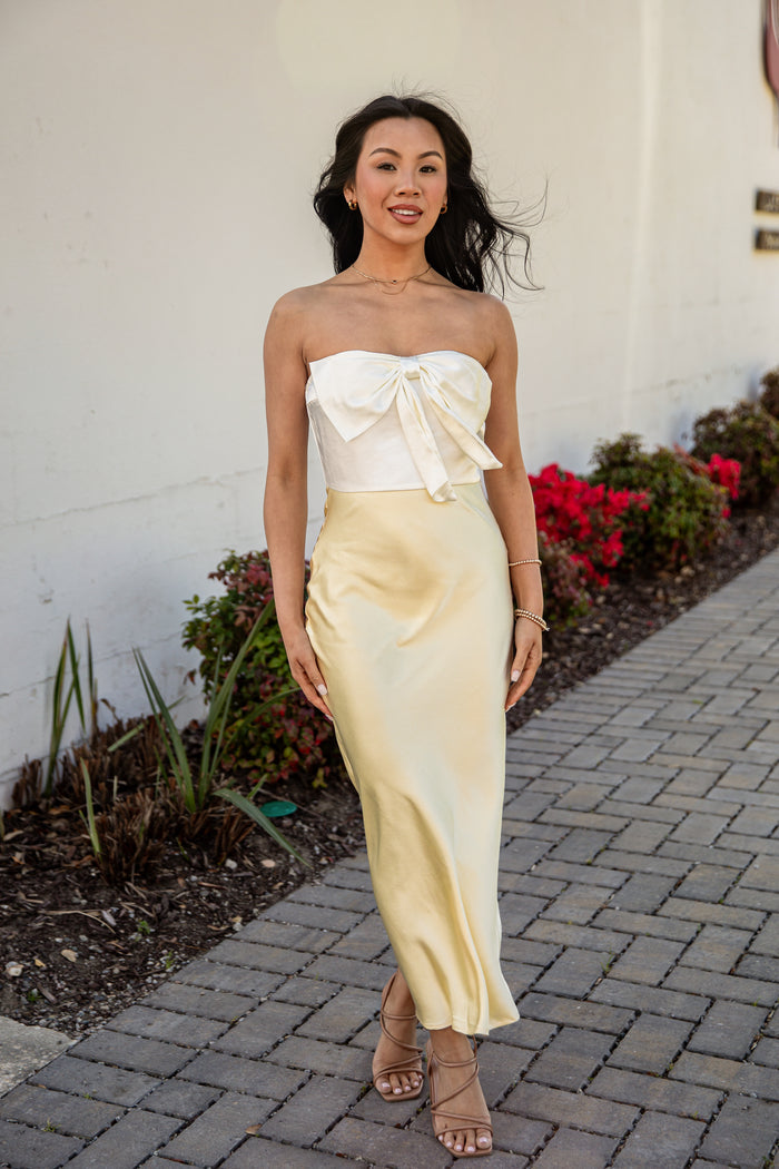 Woman in a strapless white top and yellow skirt standing on a sidewalk with plants in the background