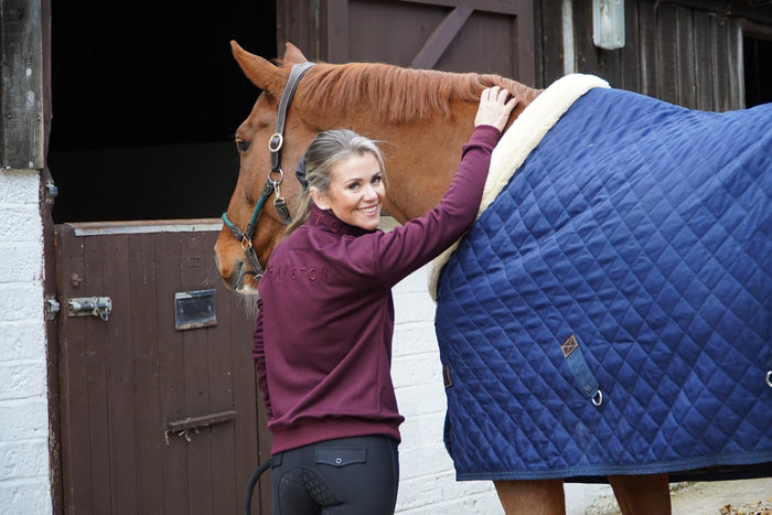 Woman preparing a horse for riding by adjusting a blue quilted saddle. highcliffe 1/4 zip jumper sweatshirt. colour match embroidery. luxury fleece lining burgundyblanket.