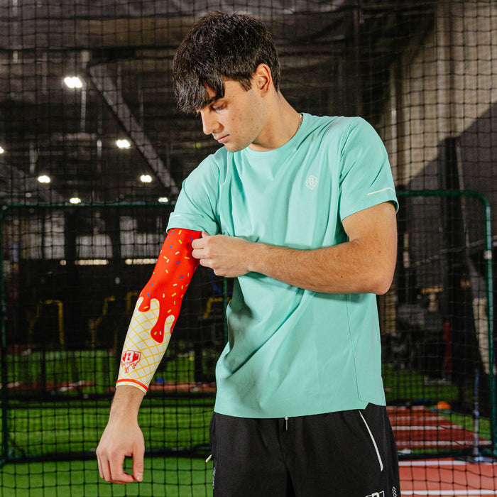 Male baseball player in a mint green shirt pulling up the ice cream arm sleeve on his left arm in an indoor batting cage