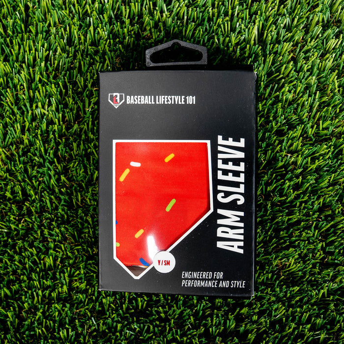 Front of the BL101 arm sleeve retail box on green turf showing the red sprinkle print through a home plate window and Y/SM size label