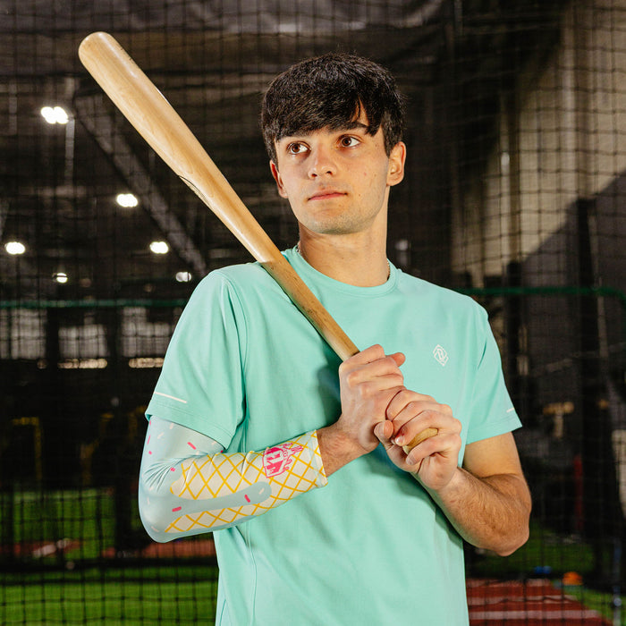 Male baseball player in a mint green shirt wearing the cotton candy ice cream arm sleeve while holding a wooden bat over his shoulder in an indoor batting cage