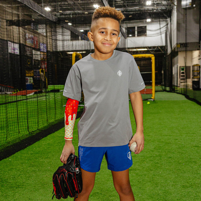 Young baseball player in a gray BL101 shirt and blue shorts wearing the ice cream arm sleeve while holding a glove and baseball in an indoor facility