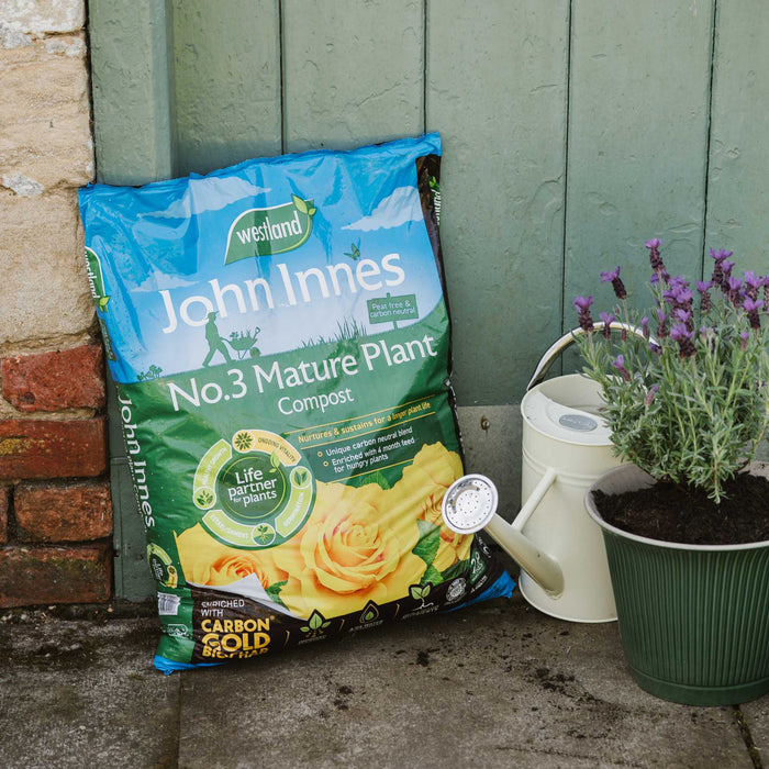 Bag of John Innes No.3 Mature Plant Compost next to a potted plant and watering can against a wooden wall.