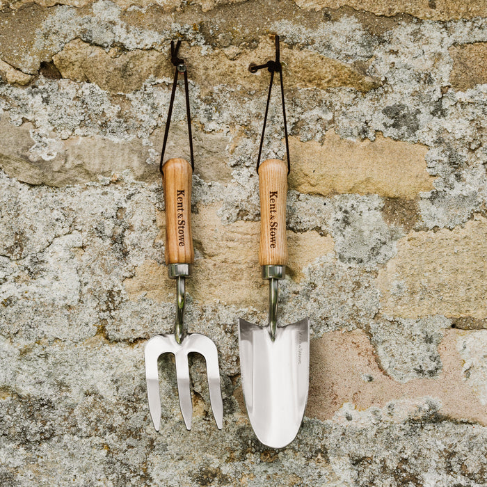 A hand trowel and hand fork with wooden handles and stainless steel heads, hanging against a stone wall.