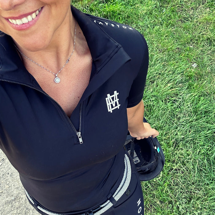 Sleek silhouette baselayer top subtle Mayston branding, maximum coverage without compromising style,  next-level performance with a bold, minimalist look. Moisture-wicking, breathable fabric with four-way stretch, it keeps you cool and comfortable. Training, Everyday or competition wear. Short Sleeve 1/4 zip with collar. classic black with grey and white branding. Woman Standing in equestrian wear