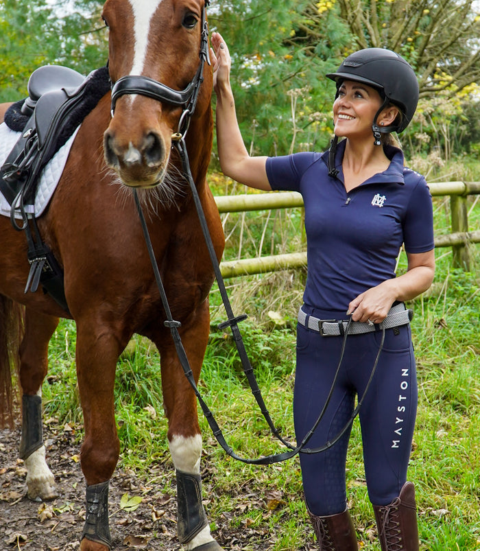 Sleek silhouette baselayer top subtle Mayston branding, maximum coverage without compromising style,  next-level performance with a bold, minimalist look. Moisture-wicking, breathable fabric with four-way stretch, it keeps you cool and comfortable. Training, Everyday or competition wear. Short Sleeve 1/4 zip with collar. navy with white branding. woman stood smiling with horse