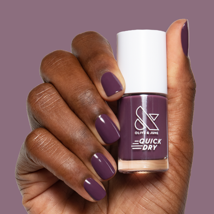 Hand with short nails painted in Plum gel polish holding Olive and June quick dry nail polish bottle against matching background
